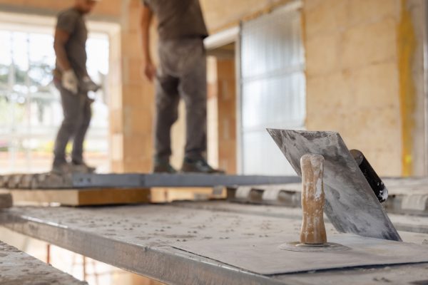 Craftsmen working inside a room standing on scaffoldings, metal trowels in the foreground. Men in grey work clothes during construction or renovation process
